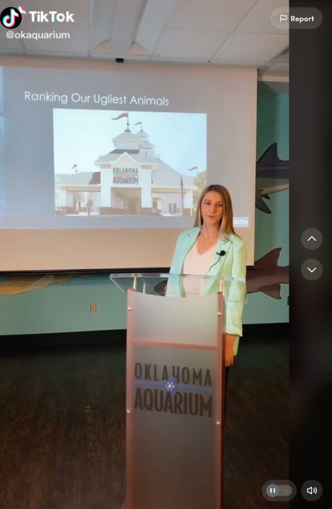 A young, blonde woman standing behind a podium that reads "Oklahoma Aquarium" and a presentation screen behind her that reads "Ranking Our Ugliest Animals" with an image of the aquarium's facility.  