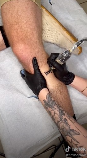 A close-up of a tattoo artist tattooing "[as]" on an Adult Swim's fan's leg